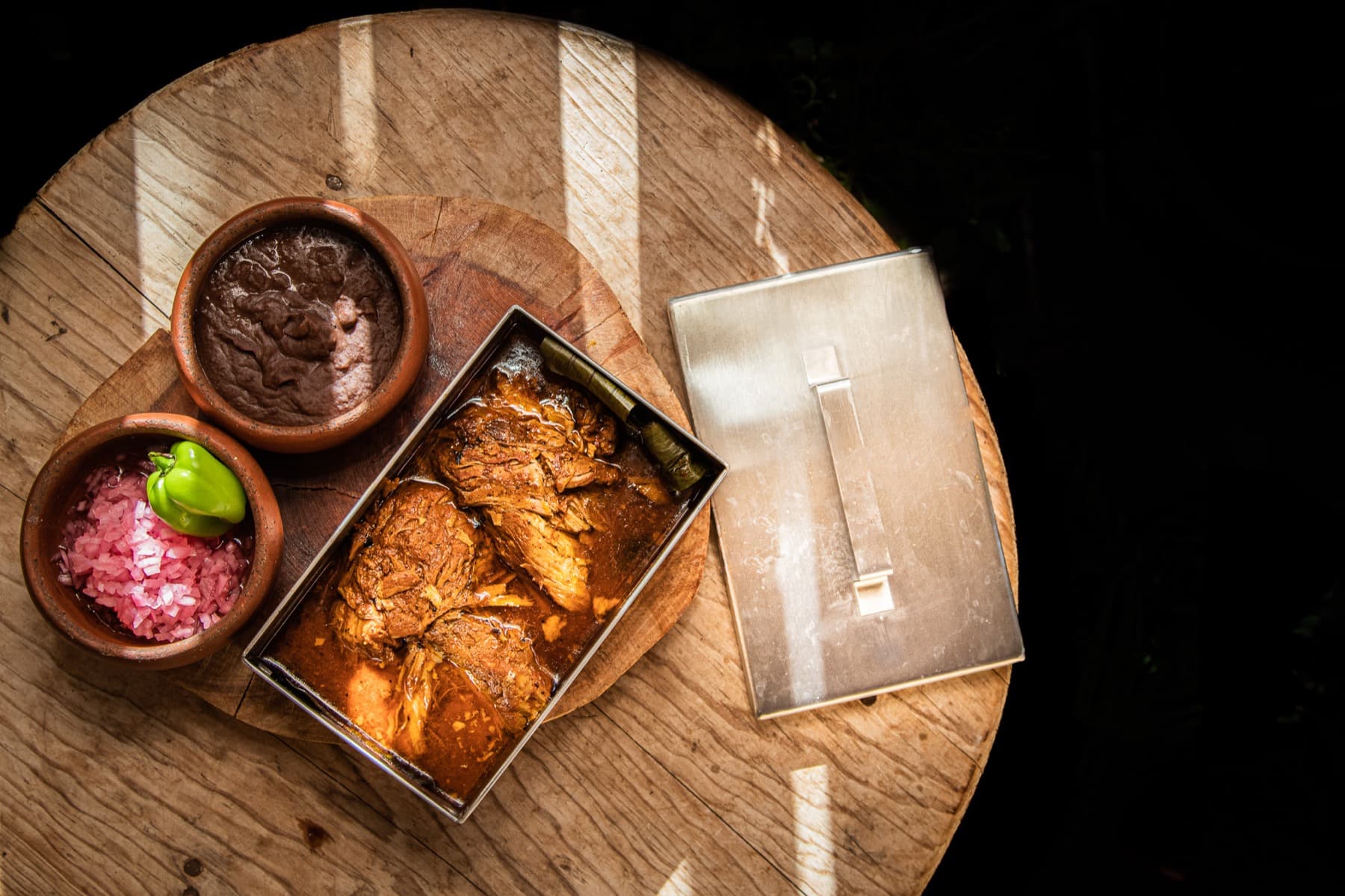 Cochinita pibil servida en caja metálica con frijol colado y cebolla morada curtida